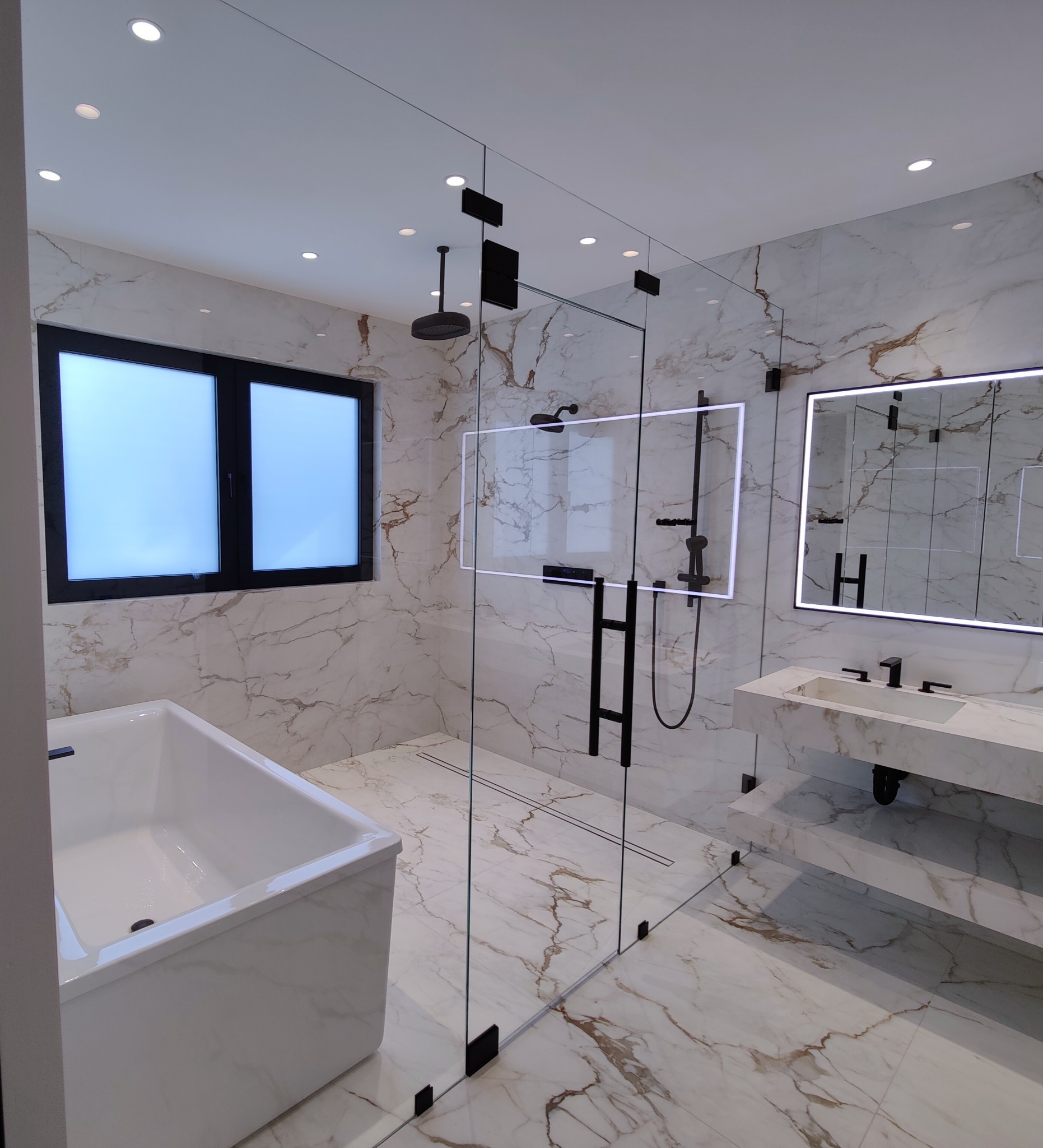 Tall clear shower enclosure with black hinges, a ladder-style pull and an overhead transom in a bathroom with veined tile.