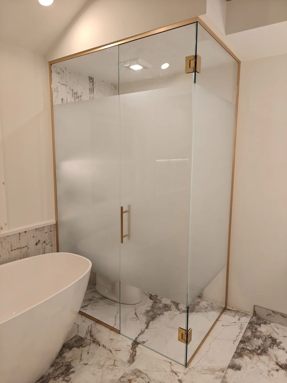 Front view of a brass-framed corner shower with frosted privacy glass, clear upper glass and marble-patterned tile.