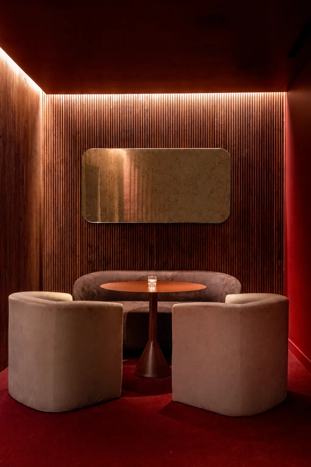 Rounded rectangular warm-toned mirror above a small table and upholstered chairs against a ribbed wood wall.