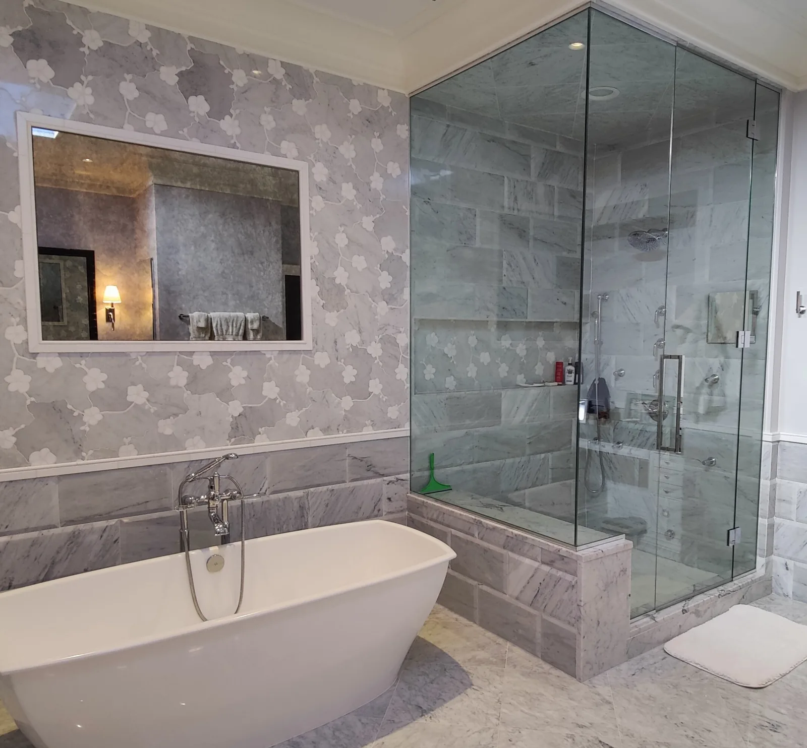 Clear ceiling-height corner shower enclosure beside a freestanding bathtub and floral wall covering.