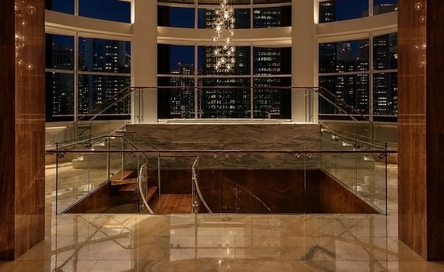 Clear glass railings around a stair opening beneath a chandelier, with wood and stone finishes and illuminated city buildings beyond tall windows.