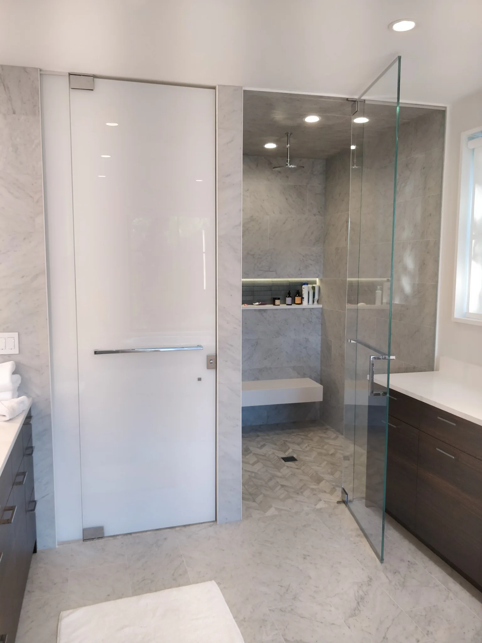 Full-height glass shower door and fixed panel in a marble bathroom with a floating shower bench