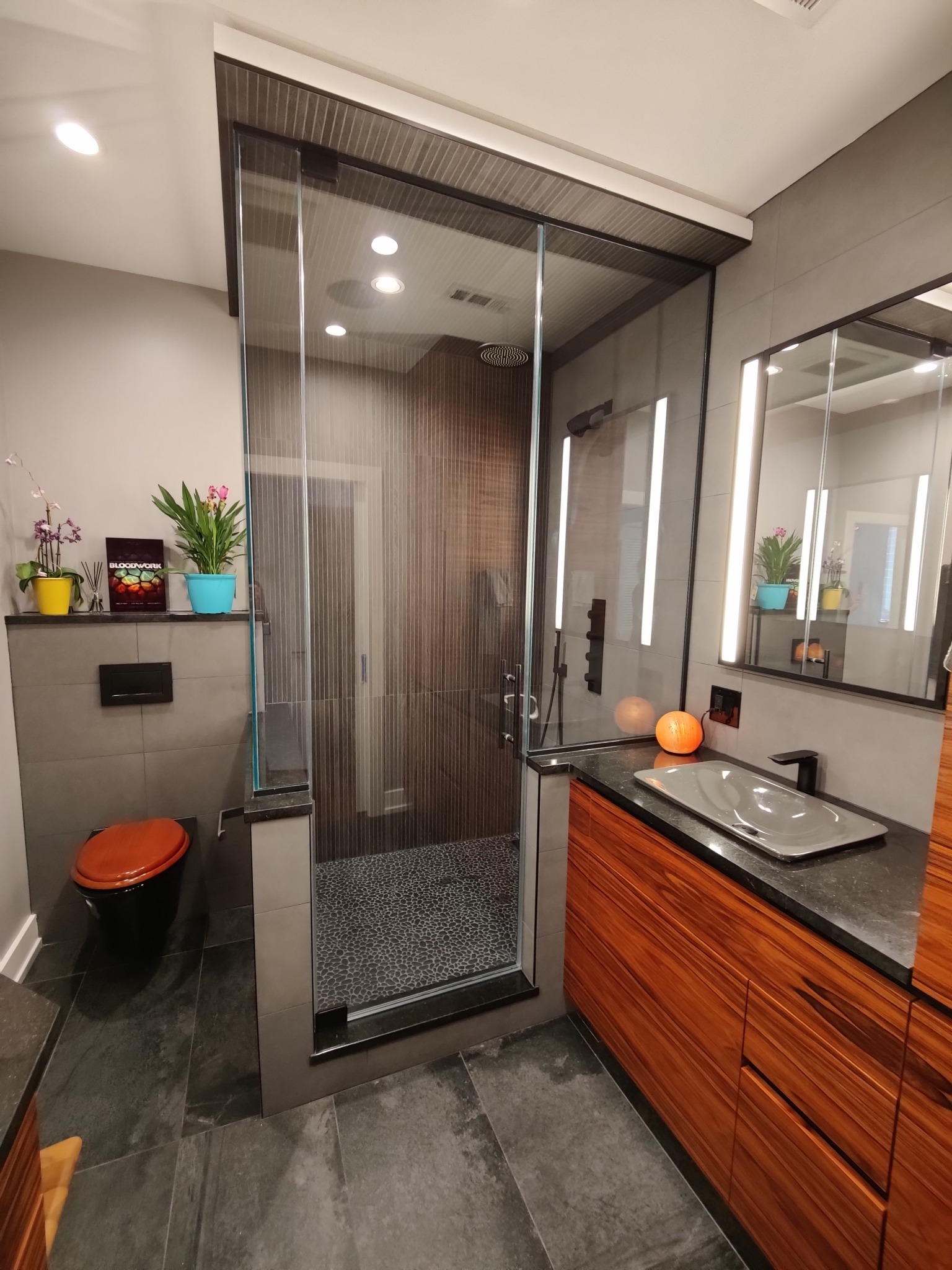 Glass shower door between fixed panels on two knee walls, beside a wood vanity and an illuminated mirror.