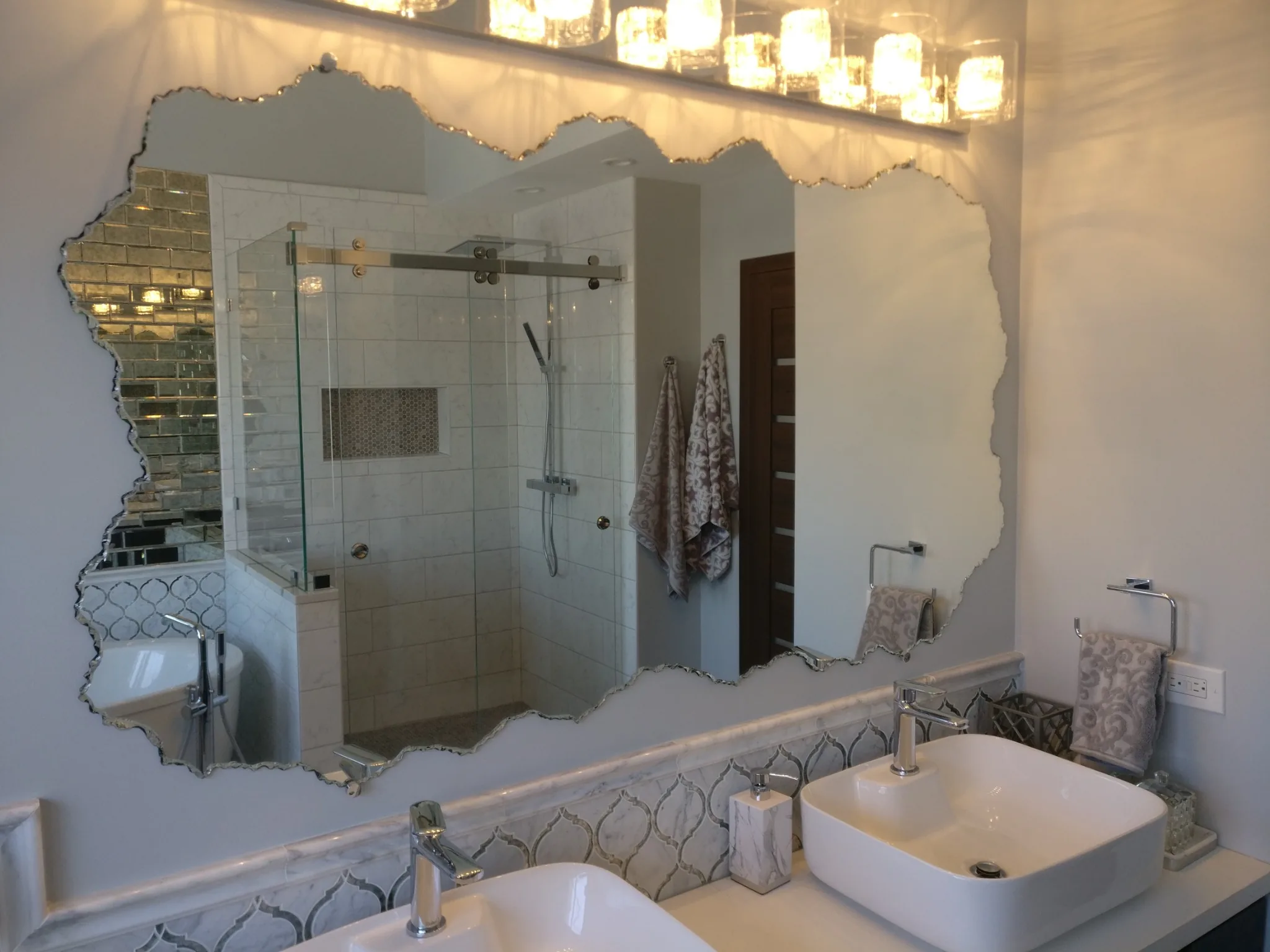 Large bathroom mirror with an irregular sculpted edge above two vessel sinks, reflecting a glass shower enclosure.