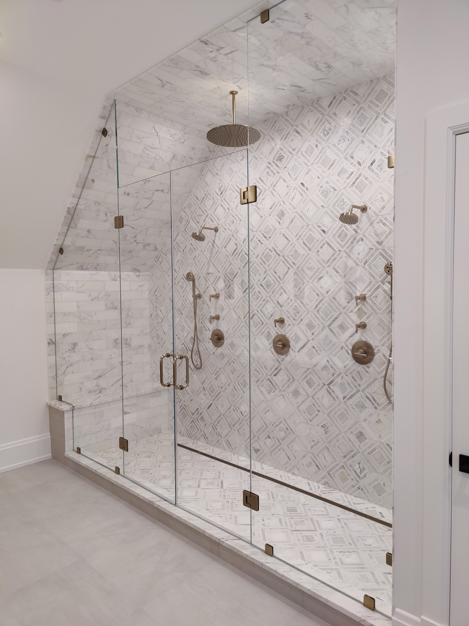 Clear shower enclosure shaped to an angled ceiling, with warm metal hinges and handles against patterned white tile.