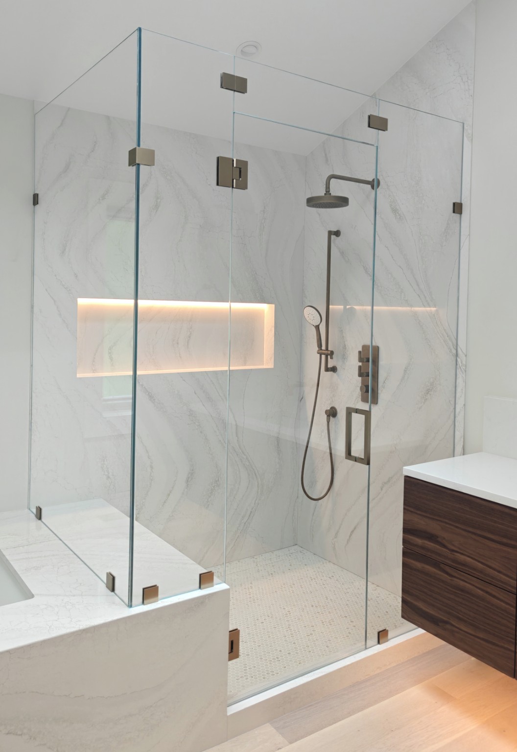 Frameless corner shower with a glass transom, warm metal hardware, marble-patterned walls and an illuminated niche beside a wood vanity.